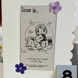 May include: A white card with a black and white illustration of a couple. The woman is sitting with her legs crossed and the man is resting his head in her lap. The text "love is... resting your head in her lap." is printed below the illustration. The card has a scalloped edge and is decorated with purple flowers.