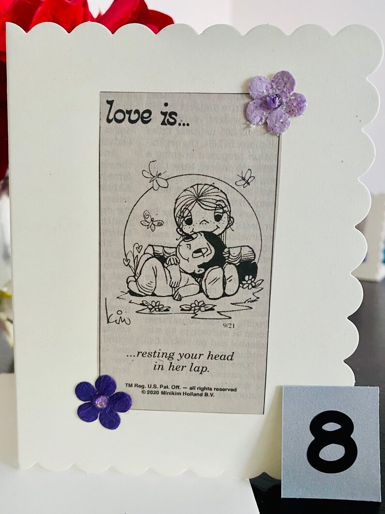May include: A white card with a black and white illustration of a couple. The woman is sitting with her legs crossed and the man is resting his head in her lap. The text "love is... resting your head in her lap." is printed below the illustration. The card has a scalloped edge and is decorated with purple flowers.