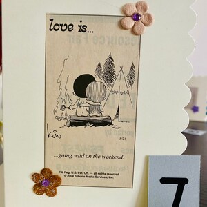 May include: A card with a cartoon illustration of a couple camping. The text on the card reads "love is... going wild on the weekend." The card is decorated with a brown flower with a purple center and a white flower with a purple center.