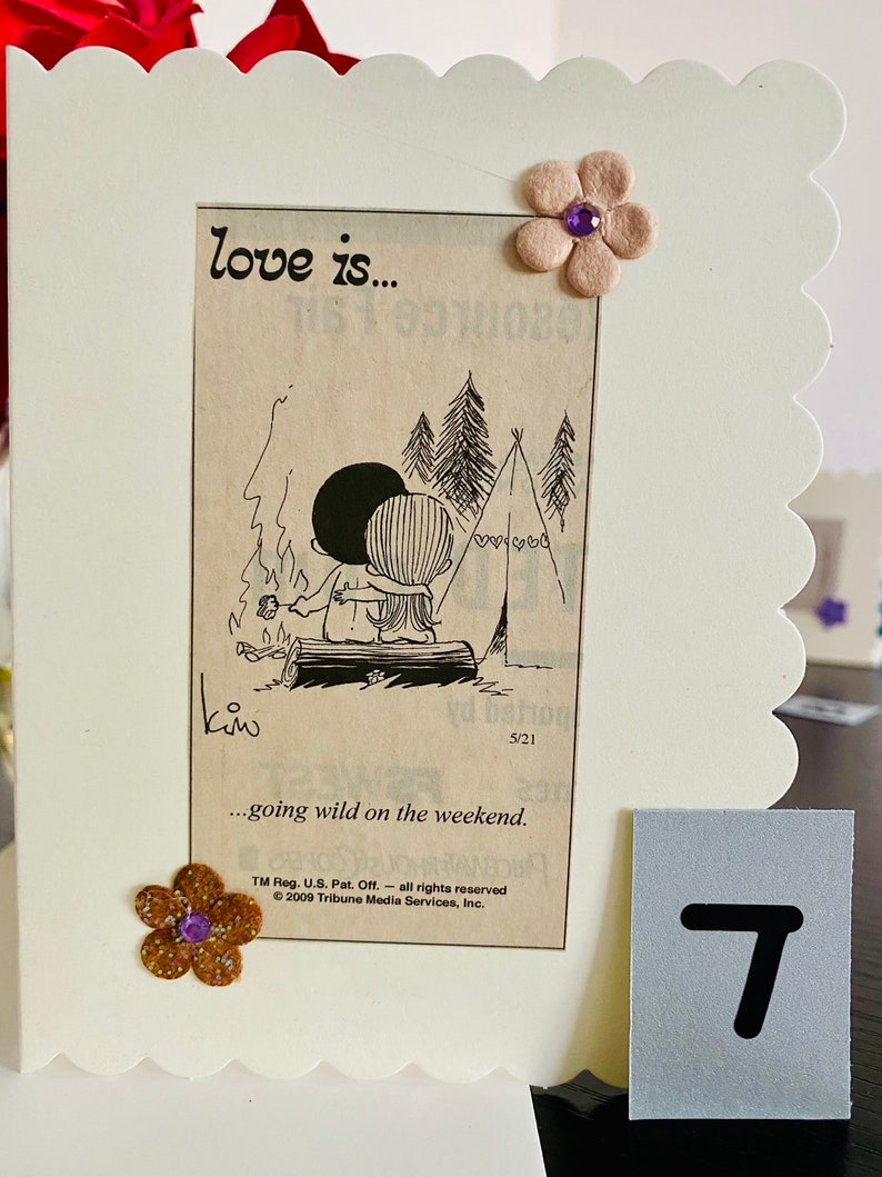 May include: A card with a cartoon illustration of a couple camping. The text on the card reads "love is... going wild on the weekend." The card is decorated with a brown flower with a purple center and a white flower with a purple center.