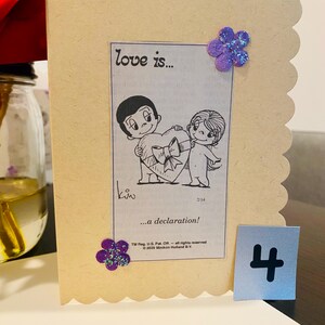 May include: A light brown card with a scalloped edge. The card features a black and white cartoon illustration of a couple holding a heart. The text "love is..." and "...a declaration!" is printed on the card. There is a purple glitter flower on the top right corner of the card. The number "4" is printed on a separate piece of paper.