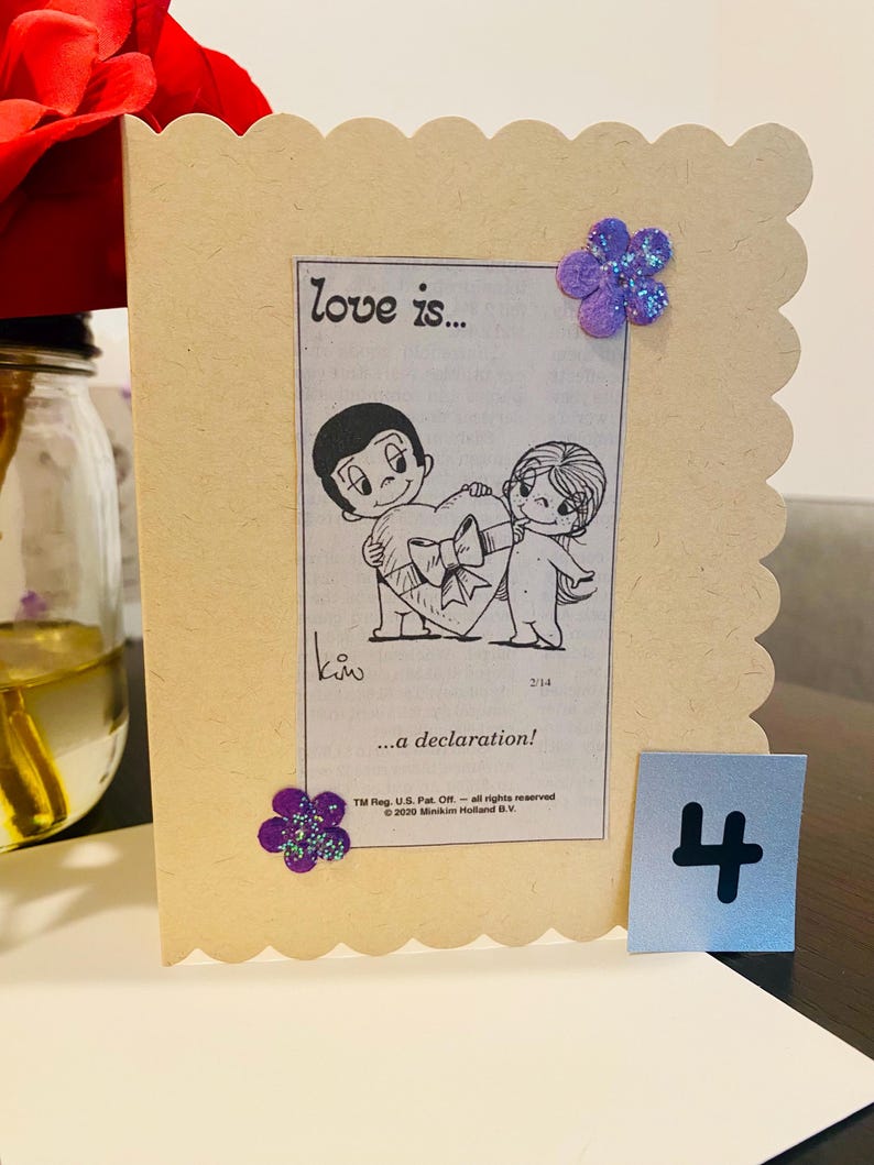 May include: A light brown card with a scalloped edge. The card features a black and white cartoon illustration of a couple holding a heart. The text "love is..." and "...a declaration!" is printed on the card. There is a purple glitter flower on the top right corner of the card. The number "4" is printed on a separate piece of paper.