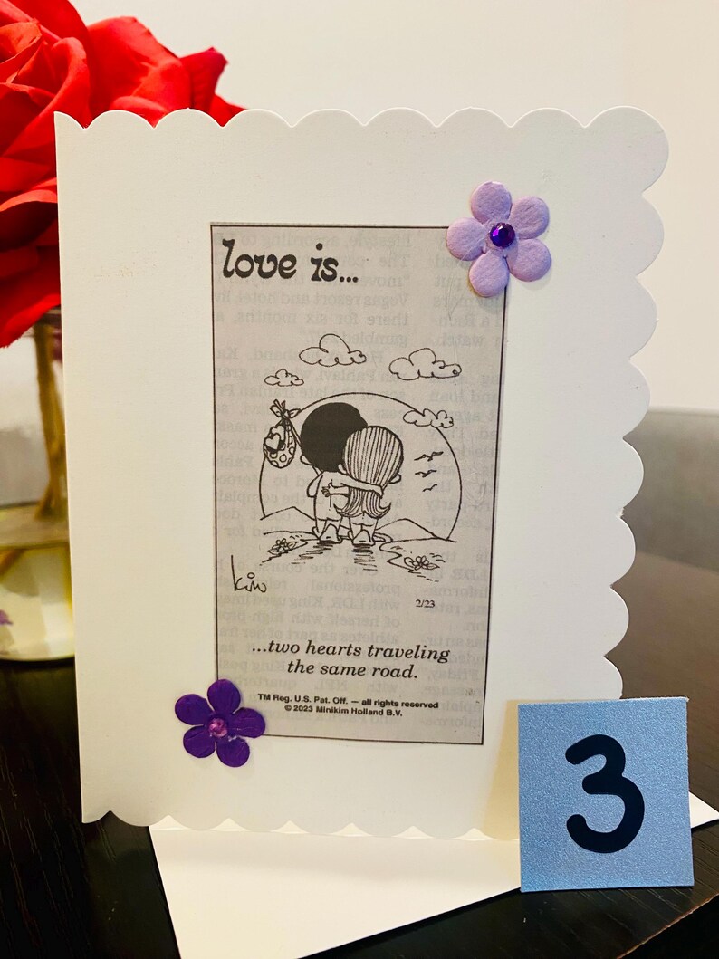 May include: A white card with a scalloped edge features a cartoon illustration of a couple holding each other. The text "love is... two hearts traveling the same road." is printed below the illustration. The card is decorated with two purple flowers.
