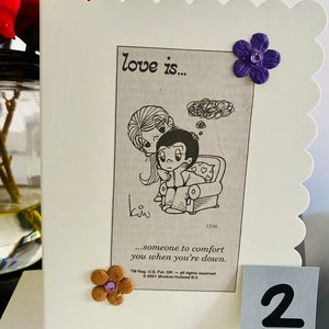 May include: A white card with a cartoon image of a couple. The card has the text "love is..." and "...someone to comfort you when you're down." The card has a purple flower and a brown flower with a purple center.