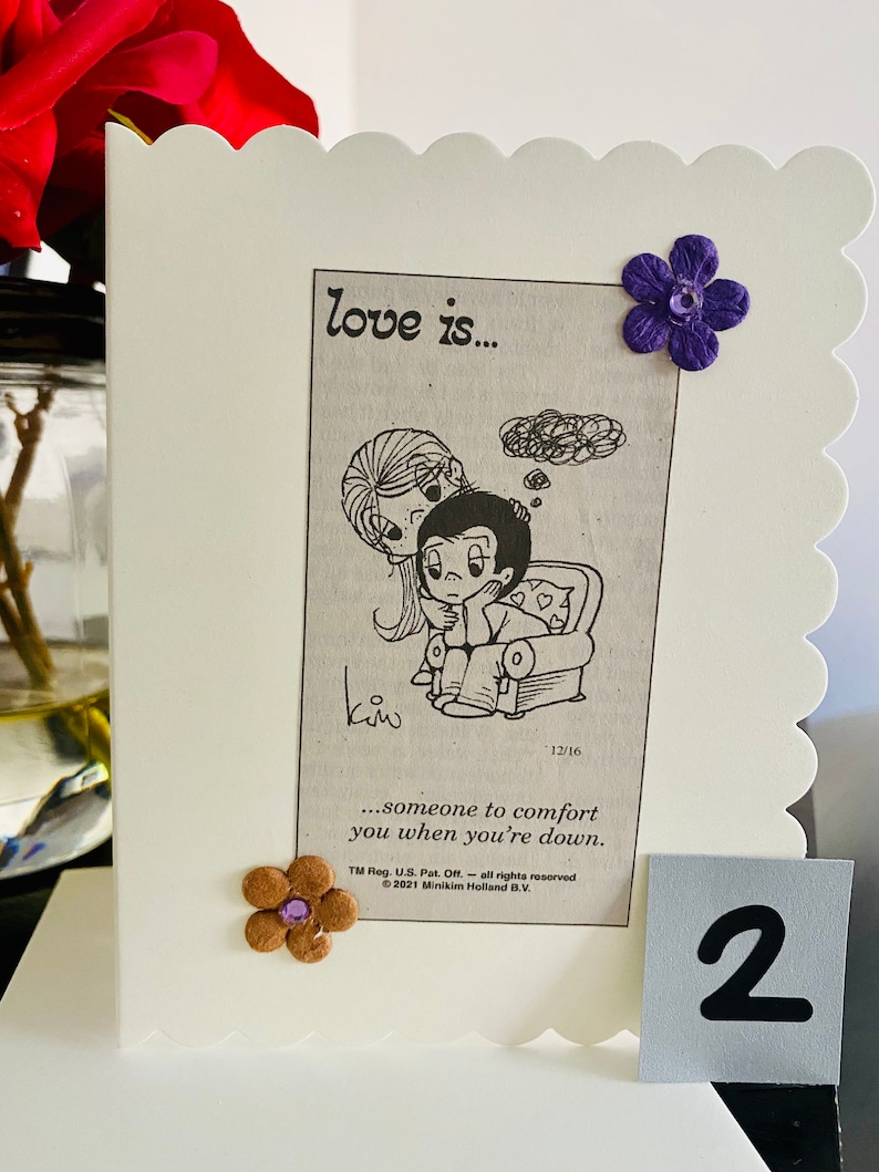 May include: A white card with a cartoon image of a couple. The card has the text "love is..." and "...someone to comfort you when you're down." The card has a purple flower and a brown flower with a purple center.