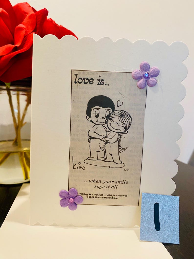 May include: A white card with a scalloped edge features a black and white cartoon illustration of a couple hugging. The text "love is..." and "...when your smile says it all." is printed on the card. The card is decorated with purple flowers.