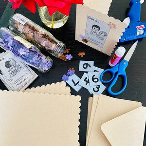 May include: A craft project with a variety of supplies including cardstock, envelopes, paper flowers, and a glue gun. The cardstock is beige and has a scalloped edge. The envelopes are beige and have a flap. The paper flowers are red and purple. The glue gun is black and has a blue handle. The text on the cardstock reads "Love is..." and "never growing too old to feel young".