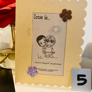 May include: A light brown card with a scalloped edge. The card features a cartoon couple with the text "love is... heart-shaped sunglasses."  The card is decorated with two brown felt flowers and a purple glitter flower.