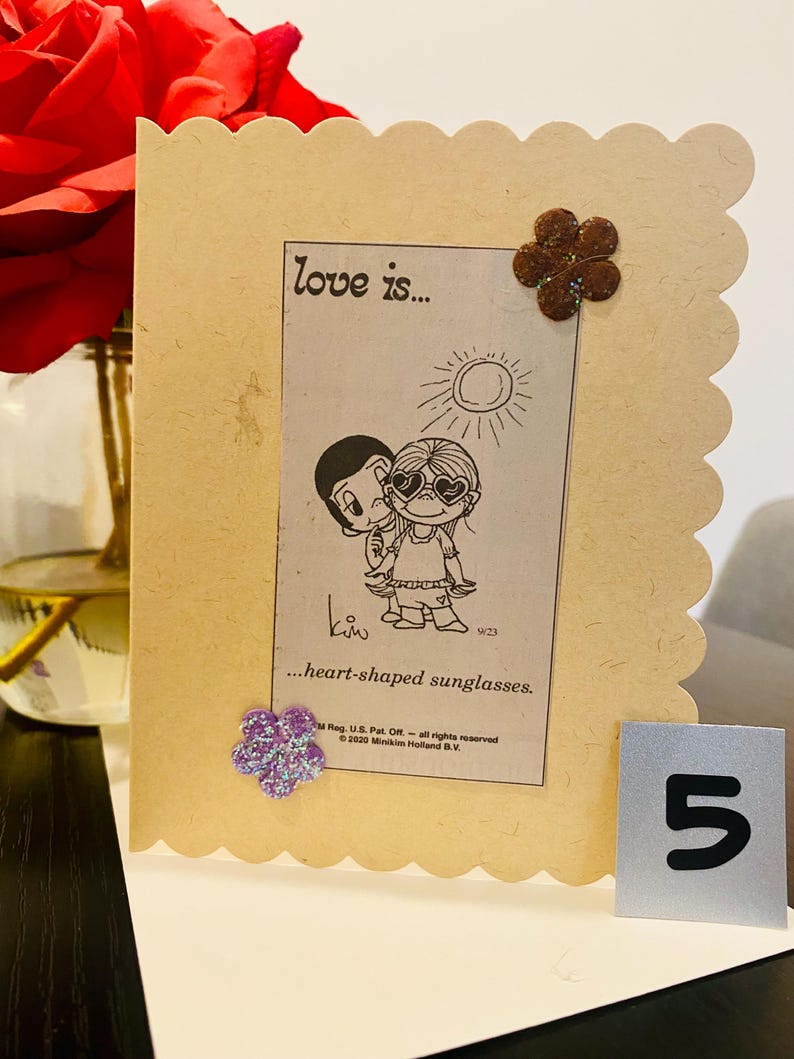 May include: A light brown card with a scalloped edge. The card features a cartoon couple with the text "love is... heart-shaped sunglasses."  The card is decorated with two brown felt flowers and a purple glitter flower.