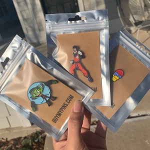 May include: Three clear plastic bags with brown cardstock inserts. Each bag contains a different enamel pin. One pin features a green cartoon character with a blue bubble, another pin features a red and blue ice cream cone, and the last pin features a cartoon character in a red and orange suit.