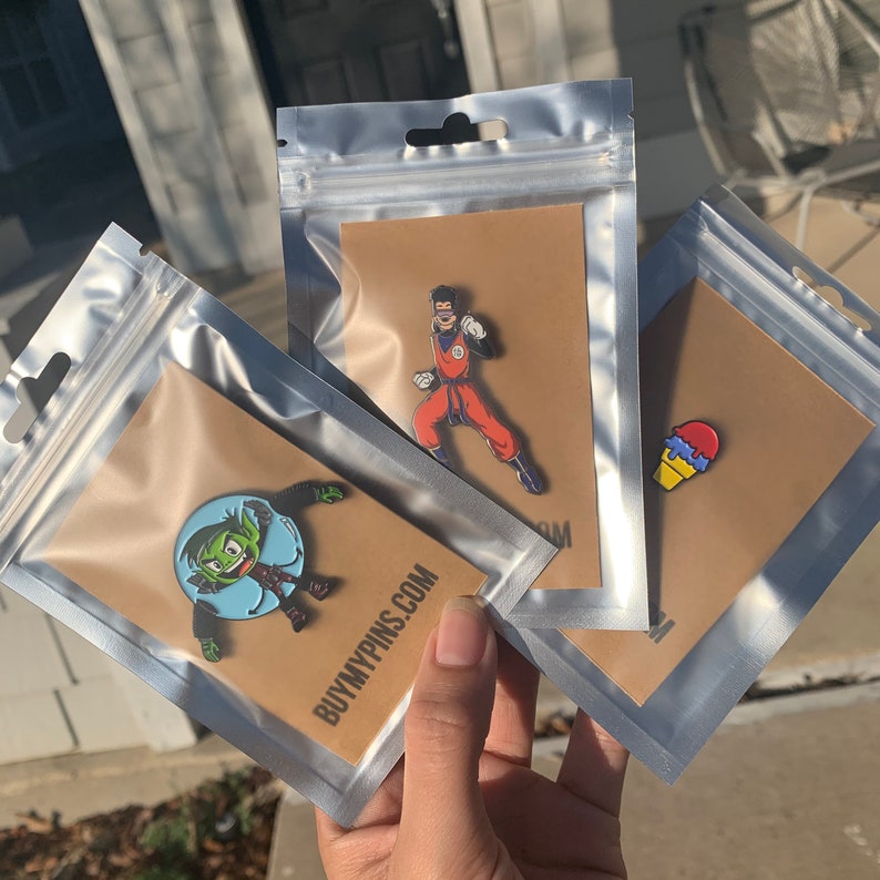 May include: Three clear plastic bags with brown cardstock inserts. Each bag contains a different enamel pin. One pin features a green cartoon character with a blue bubble, another pin features a red and blue ice cream cone, and the last pin features a cartoon character in a red and orange suit.