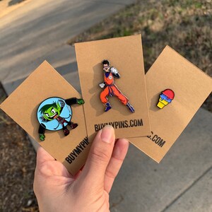 May include: Three enamel pins on brown card stock. The pins are of a green character in a bubble, a red and orange character in a fighting pose, and a blue, red, and yellow ice cream cone. The card stock has the text "BUYMYPINS.COM" printed on it.