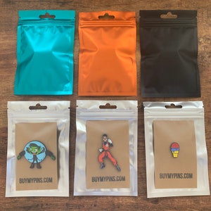May include: Three silver resealable bags with brown paper inserts. Each bag contains a different enamel pin. The first pin is green and black and depicts a cartoon character. The second pin is red, black, and yellow and depicts a cartoon character in a fighting pose. The third pin is purple, blue, and white and depicts a cartoon ice cream cone.