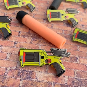 May include: A yellow and black toy gun with a red detail. The gun has a magazine with four visible bullets. The gun is a pin, and there are other similar pins in the background.