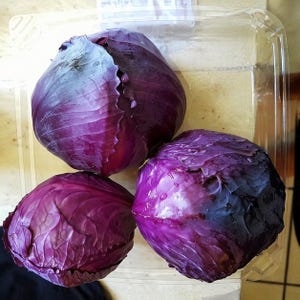 May include: Three heads of red cabbage in a clear plastic container. The cabbages are a deep purple color with textured leaves. The container sits on a light-colored surface.
