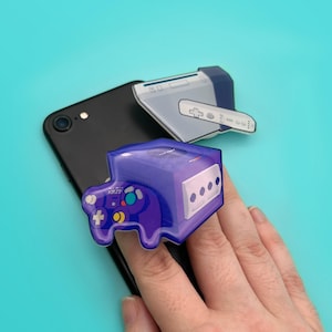 May include: A black smartphone with three colorful video game console-themed accessories. The accessories include a purple GameCube controller, a white and gray handheld console, and a white controller.