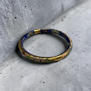 May include: A gold-toned bangle bracelet with a blue enamel band and floral designs in red, green, and white. The bracelet has a detailed, ornate appearance, suggesting a vintage or antique style. The background is a textured gray surface.