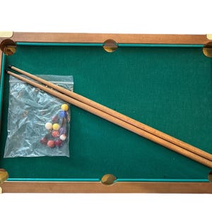 May include: A miniature pool table with a green felt surface, wooden frame, and brass corner accents. Two wooden cues and a bag of colorful pool balls are included. The table measures approximately 20 inches long.