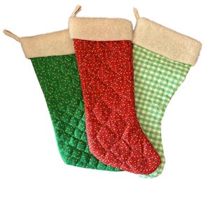 May include: Three Christmas stockings in green, red, and green and white gingham. The green stocking has a quilted pattern with white dots. The red stocking has a quilted pattern with yellow dots. Each stocking has a cream-colored faux fur cuff.
