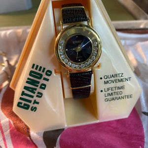 May include: A gold-tone watch with a black leather band and a black face with rhinestones around the edge. The watch is in a white and green box with the words "Gitano Studio" printed on the side. The box also states "Quartz Movement" and "Lifetime Limited Guarantee".