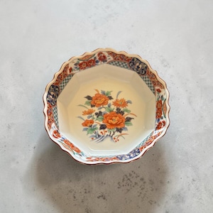 May include: An octagonal ceramic bowl with a scalloped rim, featuring a central floral motif of orange and yellow chrysanthemums with green leaves, surrounded by a decorative border of blue, red, and white geometric and floral patterns.