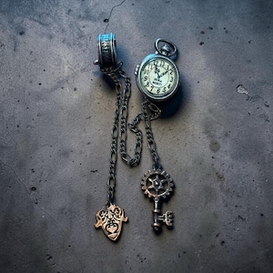 May include: A silver-toned metal accessory featuring a small round pocket watch with the text HORA MORTI on the face, connected by chains to a gear-shaped key charm and a decorative shield-shaped pendant, with a cylindrical bead at the top.