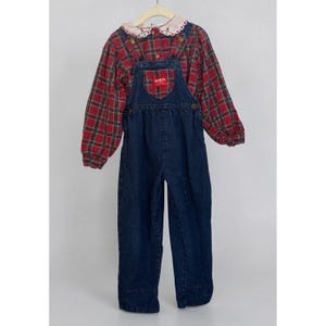 May include: A dark blue denim overall set featuring a red and green plaid long-sleeve shirt with a white collar. The collar has a floral embroidery pattern. The denim overalls have a red OshKosh B'gosh brand tag on the front chest pocket.