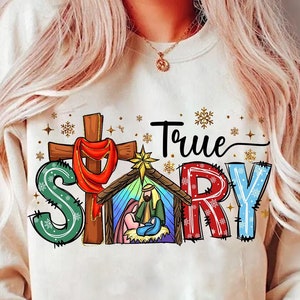 May include: A beige sweatshirt with the words "True Story" in a colorful design. The design includes a cross, a nativity scene, and snowflakes. The sweatshirt is a long-sleeved, crew neck style.