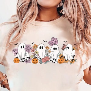 May include: A white t-shirt with a graphic of five ghosts in a line, each holding a jack-o-lantern. The ghosts are surrounded by flowers and butterflies. The shirt is a light cream color.