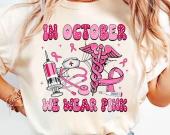 May include: Cream-colored t-shirt with a pink graphic design. The design includes the text "IN OCTOBER WE WEAR PINK" along with medical symbols such as a syringe, stethoscope, and a medical symbol. Pink ribbons are also included in the design.
