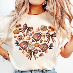 May include: Cream-colored t-shirt featuring a heart-shaped design composed of footballs, pumpkins, bows, sunflowers, and fall foliage. The design incorporates brown, orange, and yellow hues, evoking an autumn theme.