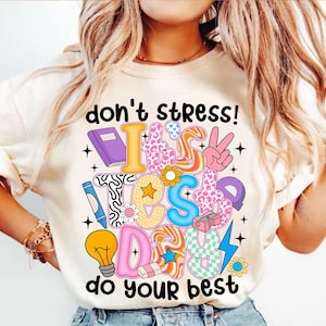 Puede incluir: Una camiseta blanca con un gráfico colorido que dice "¡no te estreses! es el día del examen haz lo mejor que puedas". El gráfico está formado por varios útiles escolares y otros diseños divertidos.