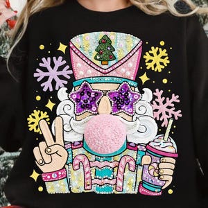 Könnte beinhalten: Schwarzes Sweatshirt mit einem skurrilen Weihnachts-Nussknacker-Design. Der Nussknacker trägt sternförmige Sonnenbrillen, eine rosa Kaugummiblase und einen festlichen Hut. Er hält einen Kaffeebecher und zeigt das Peace-Zeichen. Schneeflocken- und Sternakzente.