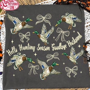 Puede incluir: Una camiseta gris oscuro con un estampado gráfico de cinco patos reales volando, varios lazos dibujados con líneas y pequeñas estrellas. El texto "Hello Hunting Season Goodbye Husband" está impreso en una fuente cursiva de color amarillo claro.