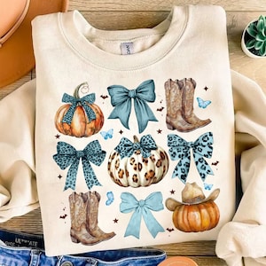 May include: Cream-colored sweatshirt featuring a fall-themed design. The design includes pumpkins, bows, cowboy boots, and butterflies. The pumpkins have leopard print and bow accents. The boots are brown, and the bows are teal.