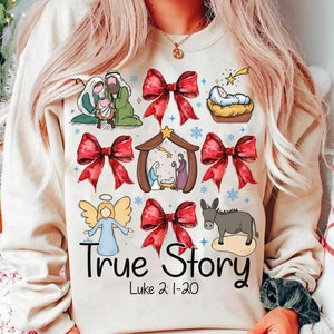 May include: Beige sweatshirt with a graphic design featuring illustrations of the Nativity scene, red bows, an angel, and a donkey. The text "True Story" and "Luke 2:1-20" are printed below the images.