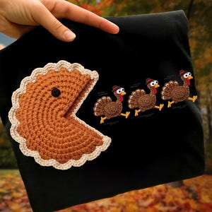 May include: Black t-shirt featuring a Pac-Man-style pie slice and three running turkeys wearing hats. The pie is brown with a tan crust and a black dot. The turkeys are brown with red wattles and black hats.