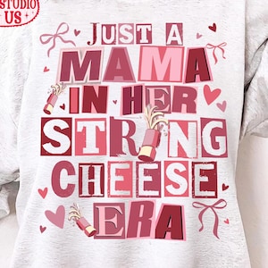 May include: A light grey sweatshirt featuring the phrase "JUST A MAMA IN HER STRONG CHEESE ERA" in pink and red lettering. The design includes hearts, bows, and lipstick illustrations. The logo "Klavia Studio US" is in the upper left corner.
