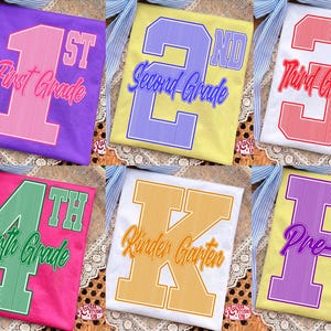 May include: A collection of six t-shirts featuring school grade designs. The shirts display large varsity-style numbers and text including 1st First Grade, 2nd Second Grade, 3rd Third Grade, 4th Fourth Grade, K Kinder Garten, and P Pre-K in various colors.