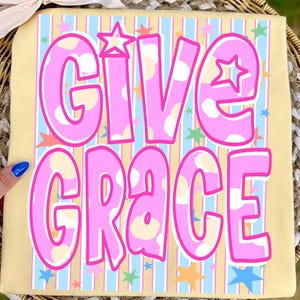 May include: A tan-colored square with the words "GIVE GRACE" in large, pink, polka-dotted letters. The background has vertical stripes in light blue and tan, with colorful stars scattered throughout. A white bow is at the top.