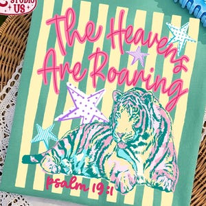 May include: A teal-colored graphic tee featuring a colorful tiger illustration with pink, teal, and yellow stripes. The text "The Heavens Are Roaring" is in pink, with star accents. The text "psalm 19:1" is at the bottom.