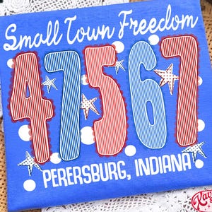 Könnte beinhalten: Blaues T-Shirt mit dem Text "Small Town Freedom" und den Zahlen "47567" in rot-blauen Streifen. Der Text "PERERSBURG, INDIANA" steht darunter, mit Sternakzenten.