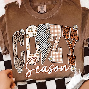 Puede incluir: Una camiseta marrón Comfort Colors con las palabras "COZY Season" en una fuente decorativa. Las letras están diseñadas con estampado de leopardo, cuadros de vichy y un patrón de tablero de ajedrez. Pequeñas margaritas acentúan el texto.