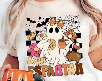 May include: Cream-colored t-shirt with a ghost graphic holding a pan dulce and a coffee cup. The ghost is surrounded by flowers, bats, and the text "Aqui Espantan" in a retro font. A person is holding an orange pumpkin mug.