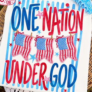May include: A white t-shirt featuring the text ONE NATION UNDER GOD in blue and red lettering. The design includes three hanging American flag banners with red and white stripes and blue star fields, accented by small red and blue stars.