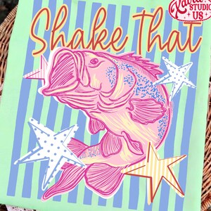 May include: A light green square with the words "Shake That" in red and yellow script. A pink fish illustration is centered, surrounded by white and yellow stars. The background has blue vertical stripes.