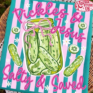 Op de afbeelding: Een turquoise en roze gestreepte keukendoek met de woorden "Pickles & Jesus Salty & Saved" in roze script. De handdoek toont een pot augurken met een roze strik en komkommerschijfjes.