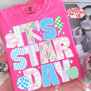 May include: A bright pink t-shirt with the text "IT'S STAR DAY" in a colorful, patterned design. The letters feature various patterns, including polka dots, floral prints, and stripes. A green and white apple graphic is also included.