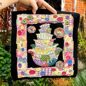 May include: A black square textile art piece with a colorful patchwork design. The artwork features a central image of stacked teacups and is framed with a patchwork border and various colored buttons.