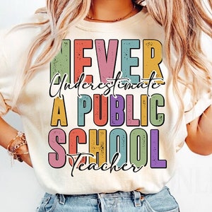 Peut inclure: Une femme portant un t-shirt beige clair avec le texte "Never Underestimate A Public School Teacher" en lettres colorées. Elle porte un jean bleu.